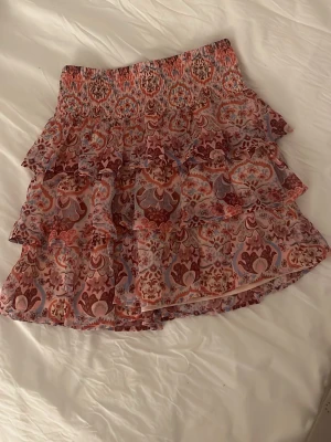 Mönstrad volangkjol i rosa och lila - Supersöt kortkjol med volanger och smock i midjan. Kjolen har ett romantiskt mönster i rosa, lila och rött och är gjord i ett lätt, luftigt material. Perfekt för dig som gillar boho-stil och vill ha något färgglatt och bekvämt.