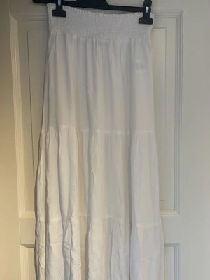 Vit långkjol med smock i midjan - Luftig och flowy vit långkjol med smockad midja och flera volanger. Perfekt för varma dagar och enkel att styla med olika toppar. Kjolen har en avslappnad passform och är tillverkad i ett lätt material.