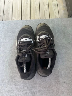 Svarta Nike sneakers med vit detalj - Säljer ett par svarta Nike sneakers med vita detaljer och klassisk swoosh på sidan. Skorna har meshmaterial på ovansidan, svarta skosnören och en låg profil. Sulan är robust och har en synlig luftkudde för extra komfort.