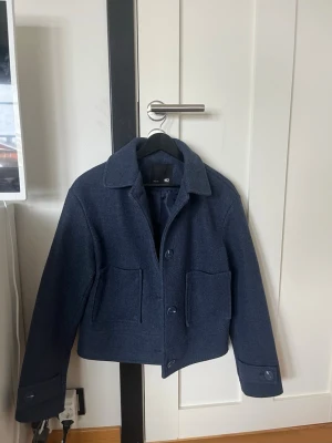 Blå kappa från Lager 157 - Snygg mörkblå jacka från Lager 157 i ullblandning. Jackan har klassisk krage, stora framfickor och knäppning med knappar framtill. Perfekt för dig som gillar stilrena och minimalistiska plagg med en boxig passform. Endast använd någon enstaka gång! 