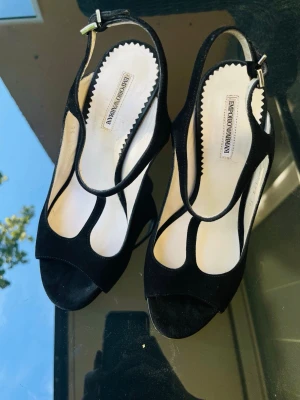Svarta pumps från Emporio Armani - Snygga svarta pumps från Emporio Armani i mocka med öppen tå och T-rem. Skorna har smal klack och justerbar rem runt hälen. Perfekta för dig som vill ha en elegant och stilren look med exklusiv känsla.