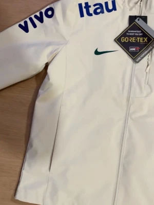 Vit Nike Brasil vindjacka GORE-TEX - Vit vindjacka från Nike med Brasil-logga och sponsormärken som Itau, Vivo och Guaraná. Jackan har huva, hel dragkedja och är tillverkad i vattentätt GORE-TEX-material. Perfekt för dig som gillar sportig stil och vill hålla dig torr. Pris kan diskuteras vid snabb affär.