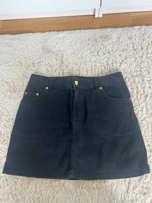 Svart kort jeanskjol  - Säljer en perfekt svart jeanskjol med guld detaljer i storlek 34! Måttet från kant till kant är 37cm och omkretsen runt är 74cm💗💗
