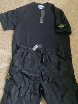 Svart set från Stone Island - Snyggt svart set från Stone Island med t-shirt och shorts. Båda plaggen har den ikoniska Stone Island-loggan på ärm och ben. T-shirten är enkel och stilren, shortsen har dragsko i midjan. Perfekt för en avslappnad och trendig look.