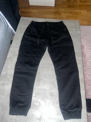 Svarta mjukisbyxor med snörning - Svarta mjukisbyxor med elastiska muddar vid bensluten och snörning i midjan. Byxorna har flera fickor och coola sömdetaljer som ger en avslappnad och trendig look. Perfekta för chill dagar eller streetwear-stil.