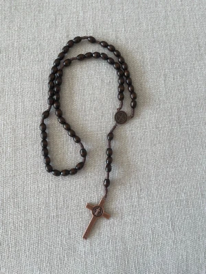 Brunt halsband med träpärlor och kors - Snyggt halsband med mörkbruna träpärlor och ett kors i trä. Halsbandet har klassisk design med runda pärlor och ett litet runt emblem ovanför korset. Perfekt för dig som gillar stilrena accessoarer med en personlig touch. Ca 45cm
