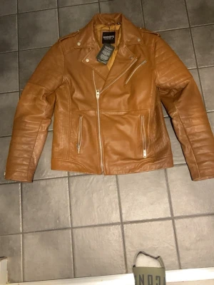 Brun skinnjacka från Barneys - Snygg brun skinnjacka från Barneys med klassisk bikerstil. Jackan har dragkedjor i silver, snedställd dragkedja framtill och flera fickor. Perfekt för dig som vill ha en stilren och tidlös look. Person i bilden är 185cm lång.