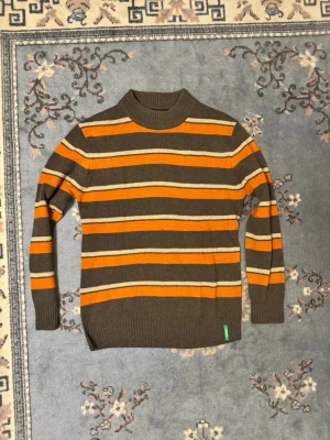 Randig brun och orange stickad tröja - Snygg stickad tröja med bruna, orangea och beigea horisontella ränder. Tröjan har hög krage och ribbstickade muddar vid ärmslut och nederkant. Perfekt för dig som gillar retrovibbar och färg. Liten grön detalj nertill på sidan.