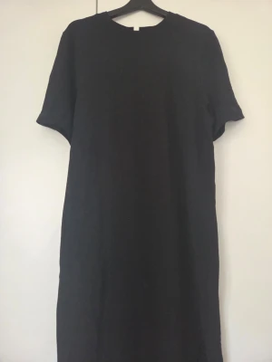 Svart långklänning med slits - Enkel svart långklänning med rund halsringning och korta ärmar. Klänningen har en rak passform och en slits framtill för extra rörelsefrihet. Perfekt för dig som gillar minimalistisk stil.