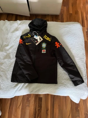  Brasilien vindjacka  - OBS ‼️‼️ Storlek M motsvarar storlek S i normal storlek , liten i storleken.                                                 Svart vindjacka från Nike med huva och dragkedja. Jackan har Brasilien-landslagets emblem, gula och orange 