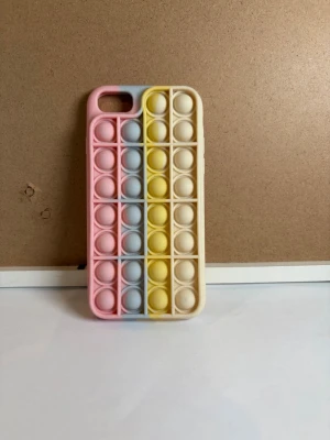 Pop it mobilskal i pastellfärger - Färgglatt mobilskal med pop it-design i pastellrosa, ljusblått, gult och beige. Skalets baksida har bubblor som kan tryckas in, perfekt för dig som gillar att pilla. Tillverkat i mjukt silikonmaterial och passar modeller från Iphone 6, 7, 8 och SE 