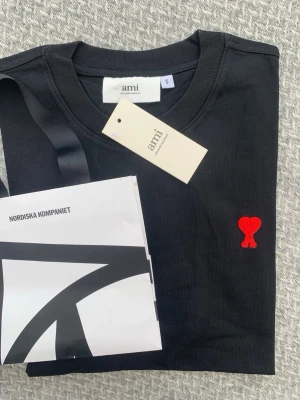 Svart t-shirt från Ami med rött hjärta - Svart t-shirt från Ami med klassisk passform och rund halsringning. På bröstet finns ett broderat rött hjärta med bokstaven A. Tillverkad i mjuk bomull, perfekt för en stilren och trendig look.