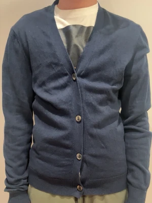 Mörkblå cardigan med knappar från pier one - En stilren mörkblå cardigan med v-ringning och fyra knappar framtill. Tröjan har långa ärmar och ribbade muddar vid ärmslut och nederkant. Perfekt att slänga över en t-shirt för en avslappnad look. Hör av er gällande frågor!!