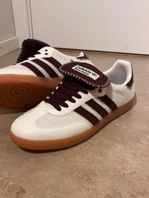 Adidas Wales Bonner Samba sneakers - Snygga Adidas Samba sneakers i samarbete med Wales Bonner. Skorna är vita med mörkbruna detaljer, klassiska tre ränder och brun gummisula. Ovandelen är i läder och mocka, med unik tunga och kontrastsömmar. Perfekta för dig som gillar stilrena och trendiga sneakers.
