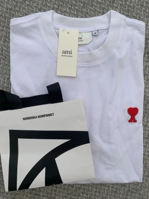 Vit t-shirt från Ami med rött hjärta - Snygg vit t-shirt från Ami Alexandre Mattiussi med ett litet rött hjärta broderat på bröstet. Klassisk rund halsringning och korta ärmar. Tillverkad i mjuk bomull för en skön känsla. Perfekt till jeans eller shorts.