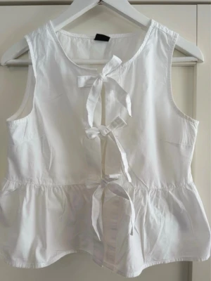 Vit ärmlös blus med knyt Gina Tricot - Supersöt vit ärmlös blus med tre knyt framtill och volang nedtill. Perfekt för en fräsch och luftig look. Blusen har rund halsringning och är tillverkad i ett lätt bomullstyg.