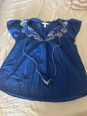 Blå broderad blus från H&M - Supersöt blå blus med vita broderier och knytband med tofsar vid halsen. Blusen har korta ärmar och en avslappnad passform, passar till både jeans och kjol.🤩❤️