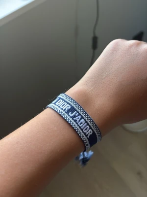 Blått Christian Dior J'Adior armband - Snyggt blått tygarmband från Christian Dior med texten 'J'ADIOR' broderad i vitt. Armbandet har justerbar knytning med tofsar i ändarna och en cool, ungdomlig vibe. Perfekt accessoar för att lyfta din outfit.