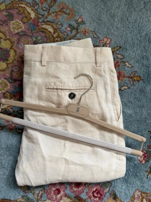 Brunello Cucinelli linnebyxor  - Snygga beige linnebyxor från Brunello Cucinelli med rak passform och klassisk design. Byxorna har bälteshällor, knappgylf och en bakficka med knapp. Perfekta för en avslappnad men stilren look. Nyskick med nypris på ca 12500kr