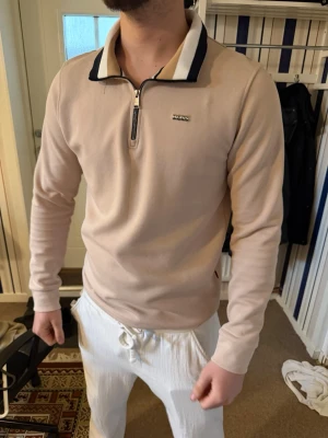 Beige långärmad tröja från Boss - Snygg beige långärmad tröja från Boss med halv dragkedja och kontrastfärgad krage i mörkblått. Tröjan har en liten Boss-logga på bröstet och är tillverkad i ett mjukt, bekvämt material. Perfekt för en clean och stilren look. Nypris: 2000.