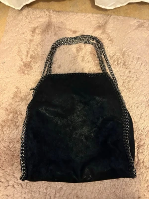 Svart shoppingväska med kedjedetaljer - Cool svart shoppingväska med silvriga kedjor längs kanterna och som handtag. Väskan har en mjuk, något glansig yta och en rymlig form som rymmer mycket. Perfekt för dig som vill ha en trendig och praktisk väska till vardags.