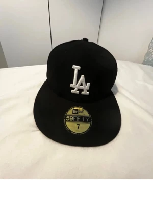 Svart LA New Era 59FIFTY keps - Säljer en svart New Era 59FIFTY keps med LA-logga broderad i vitt framtill. Klassisk flat peak och strukturerad passform. Kepsen är tillverkad i ett slitstarkt material och har officiella MLB-märken på insidan.