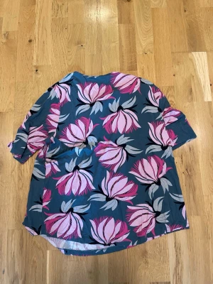 Alice Bizous Blommig skjorta stl xl i blått och rosa - Säljer en skjorta med stort blommigt mönster i rosa och grå toner på blå botten. Skjortan har korta ärmar och avslappnad passform, perfekt för dig som gillar färgstarka plagg. Materialet känns lätt och luftigt, troligen bomull eller polyester.