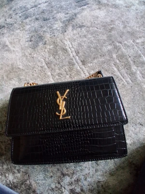 Svart axelväska från Yves Saint Laurent - Snygg svart axelväska från Yves Saint Laurent med krokodilpräglat läder och guldfärgad kedja. Väskan har YSL-logga i guld på framsidan och stilren, rektangulär form. Perfekt för dig som gillar lyxiga accessoarer och trendig design.