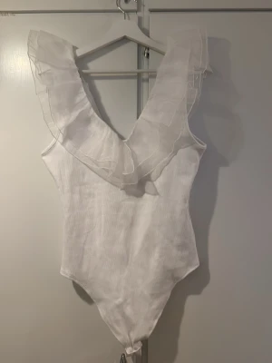 Vit body med volang från MISU - Säljer en snygg vit body från MISU med djup V-ringning och stora volangdetaljer över axlarna. Tillverkad i ett lätt, tunt tyg som ger en luftig känsla. Perfekt för dig som vill ha något extra till outfiten!