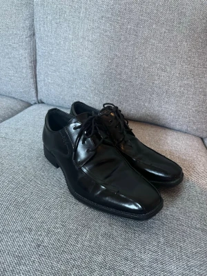 Svarta finskor i skinn - Stilrena svarta loafers i blankt skinn med snörning och fyrkantig tå. Perfekta för dig som gillar en clean och tidlös look. Skorna har diskreta sömmar och låg klack. Passar till både dressade och avslappnade outfits.