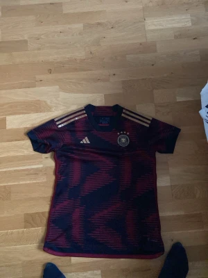 Tysklands matchtröja Adidas M - Säljer en Tysklands fotbollströja från Adidas i storlek M. Tröjan är mörkblå med röda mönster och har guldiga ränder på axlarna samt Tysklands landslagsmärke på bröstet. Regular fit och tillverkad i funktionsmaterial.