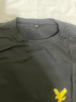 Svart topp från Lyle & Scott - Svart topp från Lyle & Scott med rund hals och korta ärmar. På bröstet finns den klassiska gula örnloggan broderad. Tillverkad i mjuk bomull för en bekväm känsla och stilren look.
