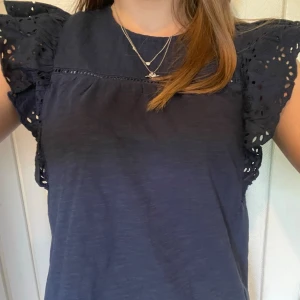 Mörkblå blus med volangärmar -  mörkblå blus med broderade volangärmar och rund hals. Luftigt material och fina detaljer vid axlarna gör den perfekt till jeans eller kjol. 
