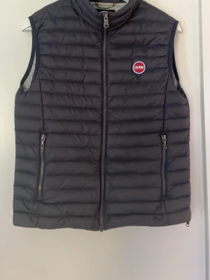 Svart dunväst från Colmar Originals - Snygg mörk blå dunväst från Colmar Originals med quiltad design och två dragkedjefickor framtill. Västen har Colmar-logga på bröstet. Perfekt att slänga över en hoodie eller tröja för extra värme. Klassisk och stilren look.