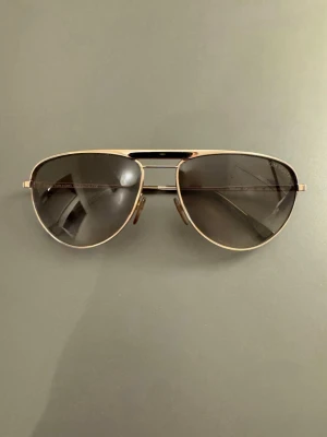 Tom Ford William Solglasögon TF207 28F - Tom Ford solglasögon i pilotmodell (modell ”William”). Bågarna är i glänsande roséguld/ljusguld med tonade bruna glas.  Glasögonen: I gott använt skick! Glasen är klara och fina, ramen är rak och stabil, och gångjärnen är fasta. De ikoniska ”T”-logotyperna på sidorna är blanka och rena.  Fodralet: Medföljer gör ett original Tom Ford sammetsfodral. Observera att fodralets material har åldrats och blivit slitet/fläckigt på både ut- och insidan (se bilder). 