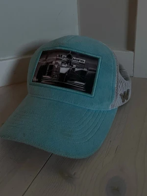 Ljusblå keps från Wonti med F1-motiv - Snygg turkos velvetkeps från Wonti med ett svartvitt F1-motiv framtill. Kepsen har böjd skärm, mesh på sidorna och justerbar baksida. Perfekt accessoar för motorsportfans och till sommaren som vill sticka ut med en unik stil. Pris kan diskuteras vid snabb affär. Den har några fläckar på den men de går att tvätta bort