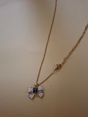 Guldhalsband med vit rosettberlock - Sött halsband med tunn guldfärgad kedja och en vit rosettberlock. Rosetten har en liten dekorativ detalj i mitten som ger ett unikt uttryck. Perfekt accessoar för att lyfta din outfit med en lekfull touch.