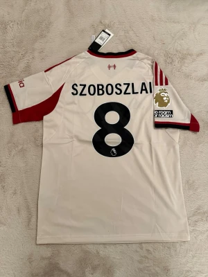 Liverpool bortatröja Szoboszlai #8 - Snygg vit Liverpool FC bortatröja från Adidas med röda detaljer och svarta inslag. Tröjan har Szoboszlai och nummer 8 på ryggen, Premier League-märke på ärmen och klubbmärke samt sponsorlogga på bröstet. Tillverkad i lätt och ventilerande material.