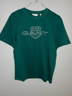 Grön GANT t-shirt med logga - Snygg grön t-shirt från GANT med stor vit logga och sköldtryck på bröstet. Klassisk rund halsringning och korta ärmar. Tillverkad i mjuk bomull som känns skön mot huden. Perfekt för en avslappnad och stilren look.