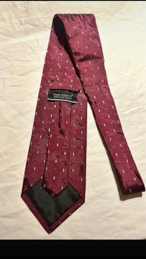 Vinröd slips från Peter Adams - Säljer en vinröd slips från Peter Adams med diskret mönster i vitt och rosa. Slipsen har klassisk form och är tillverkad i ett glansigt material som ger en elegant look. Perfekt för att lyfta din outfit med lite extra färg.