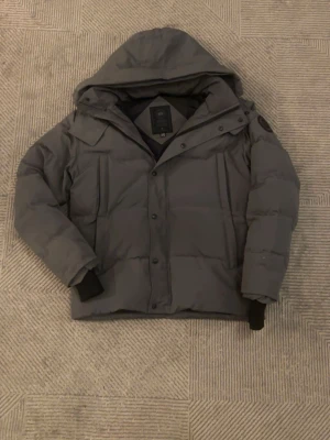 Canada goose wyndham parka jacket - Snygg grå pufferjacka med stor huva och raka linjer från Canada goose. Fur och väskan följs med också! Det finns en skada vid högra fickan, men jag har sytt den så den är lik stark som förut!! Jackan har två stora bröstfickor, muddar vid ärmsluten och en patch på ena ärmen. Perfekt för kalla vinterdagar och har en modern, clean look.