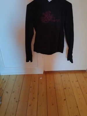 Svart hoodie med rosa tryck - Svart hoodie med huva och lång ärm. Framtill finns ett rosa grafiskt tryck och insidan av huvan är fodrad i rosa tyg. Hoodien har en klassisk passform och är tillverkad i mjukt material, perfekt för en avslappnad stil.