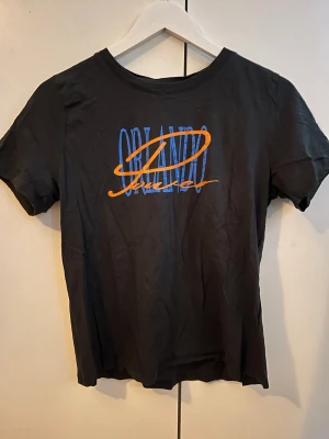 Svart Orlando Magic t-shirt - Cool svart t-shirt med tryck i blått och orange där det står 'Orlando' och 'Magic' på bröstet. Klassisk rund hals och korta ärmar. Perfekt för dig som gillar sportiga och avslappnade looks.