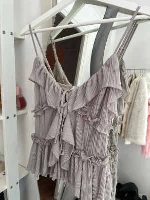 Ljusgrå volangig linnetopp - Supersöt ljusgrå linnetopp med tunna axelband och volangdetaljer. Toppen har ribbad struktur, omlottkänsla framtill och knyt i mitten. Perfekt för en trendig och luftig look.