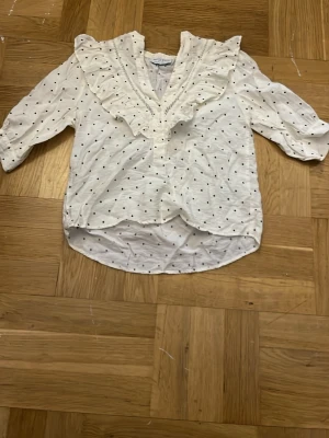 Vit blus med volang och prickar - Söt vit blus med svarta små spader på och bred volang längs v-ringningen. Blusen har trekvartsärm. 