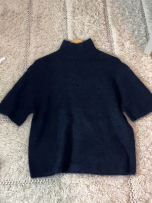 Svart stickad tröja med polokrage - Mysig svart stickad tröja med hög polokrage och korta ärmar. Tröjan har en avslappnad passform och är perfekt för lager på lager. Materialet ser ut att vara ull eller en ullmix, vilket gör den både varm och stilren.