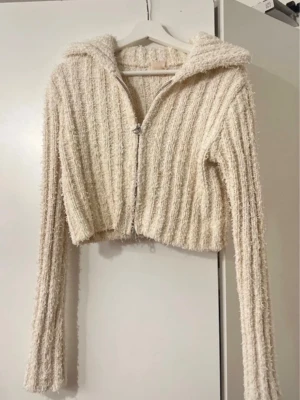 Beige fluffig cropped hoodie - Mysig beige hoodie i fluffigt, ribbstickat material. Croppad modell med dragkedja framtill och långa ärmar. Perfekt för dig som gillar en avslappnad och trendig stil