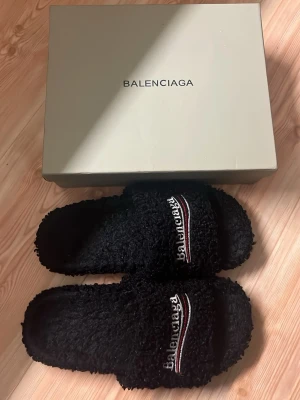 Fur slides - Säljer ett par svarta, fluffiga tofflor med broderad logga och rödvit detalj på ovandelen. Tofflorna har öppen tå och är tillverkade i mjukt, teddyliknande material för extra komfort. Perfekta för att chilla hemma med stil. STORLEK 44 SMÅ LITE SMÅ I STORLEK.