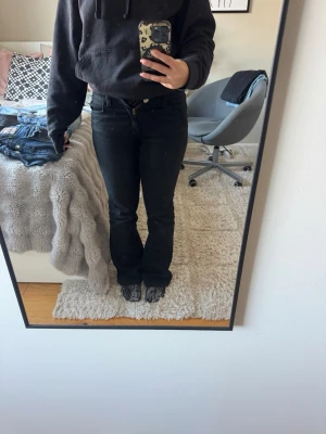 Svarta midwaist bootcut jeans - Säljer ett par svarta bootcut jeans med medelhög midja och knappgylf med två knappar. Jeansen har en klassisk femficksmodell och är tillverkade i ett stretchigt denimtyg som sitter snyggt på benen. Säljer pga att de inte passar längre 