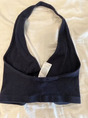 Mörkblå ribbad halterneck topp - Snygg mörkblå ribbad halterneck topp med knappar framtill. Toppen är croppad och har djup v-ringning, perfekt för en trendig och avslappnad stil. Materialet är mjukt och stretchigt för extra komfort.